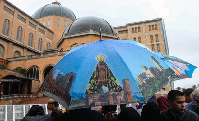 Com o Santuário Nacional de Nossa Senhora Aparecida ao fundo, fiéis aguardam na chuva a passagem do Papa Francisco em Aparecida, SP